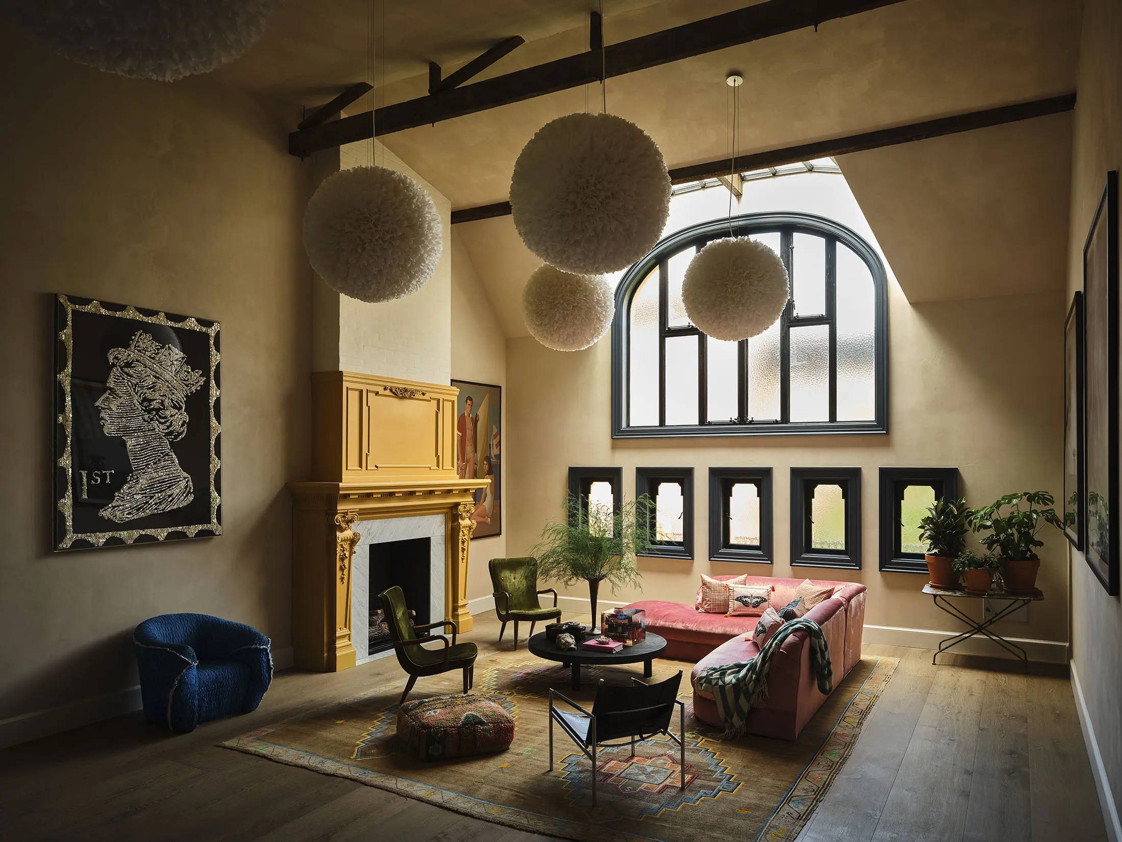 Spacious, stylish living room with high ceiling and large arched window. Features a grand yellow fireplace, pink sofa, and green chairs on a patterned rug. Unique spherical pendant lights hang from exposed beams. Warm lighting enhances cozy ambiance and rich textures.