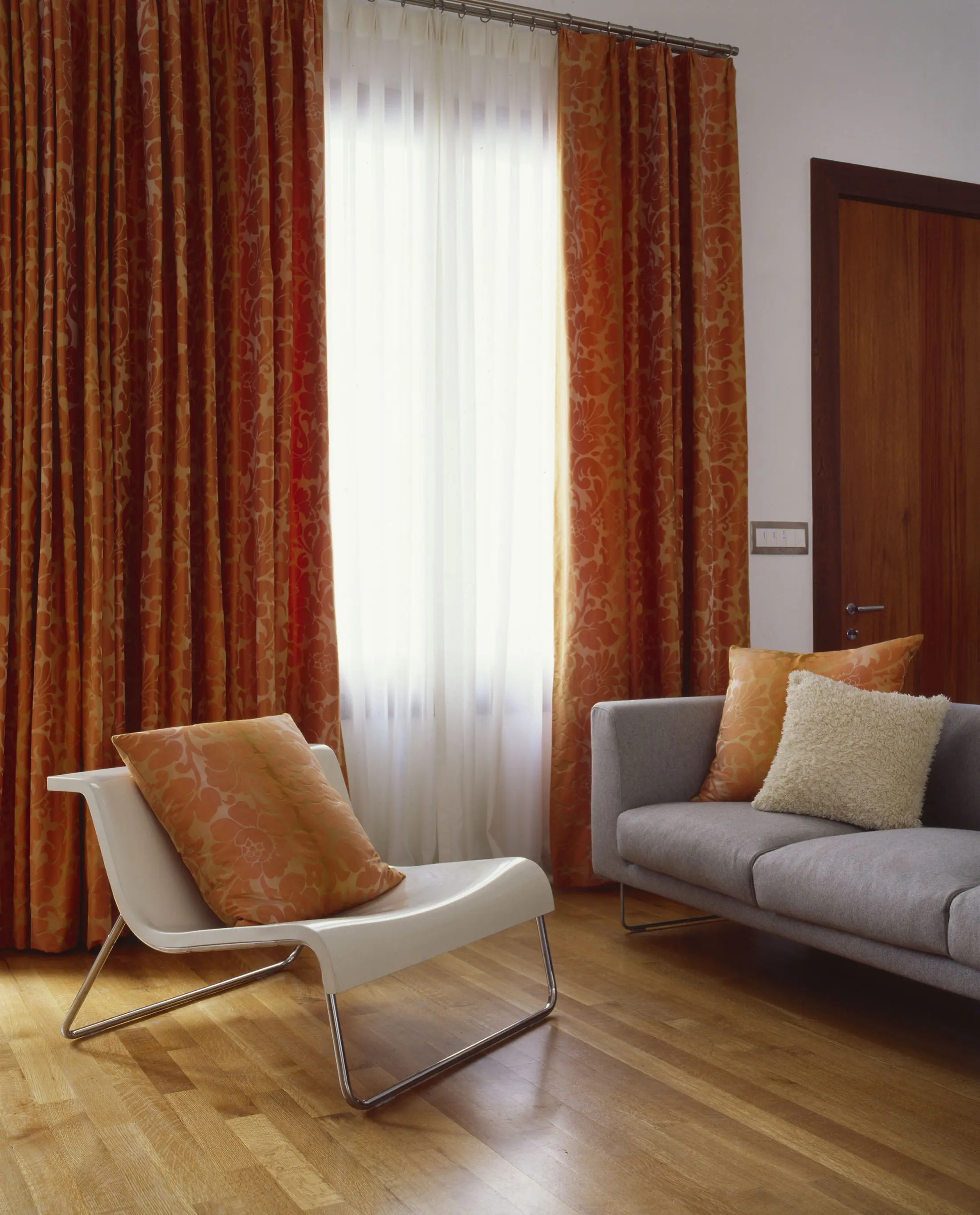 A modern living room with a grey sofa adorned with a white cushion, next to a sleek white lounge chair with orange floral pillows. The room features a wooden floor and is illuminated by natural light coming through white sheer curtains and vibrant orange patterned drapes.
