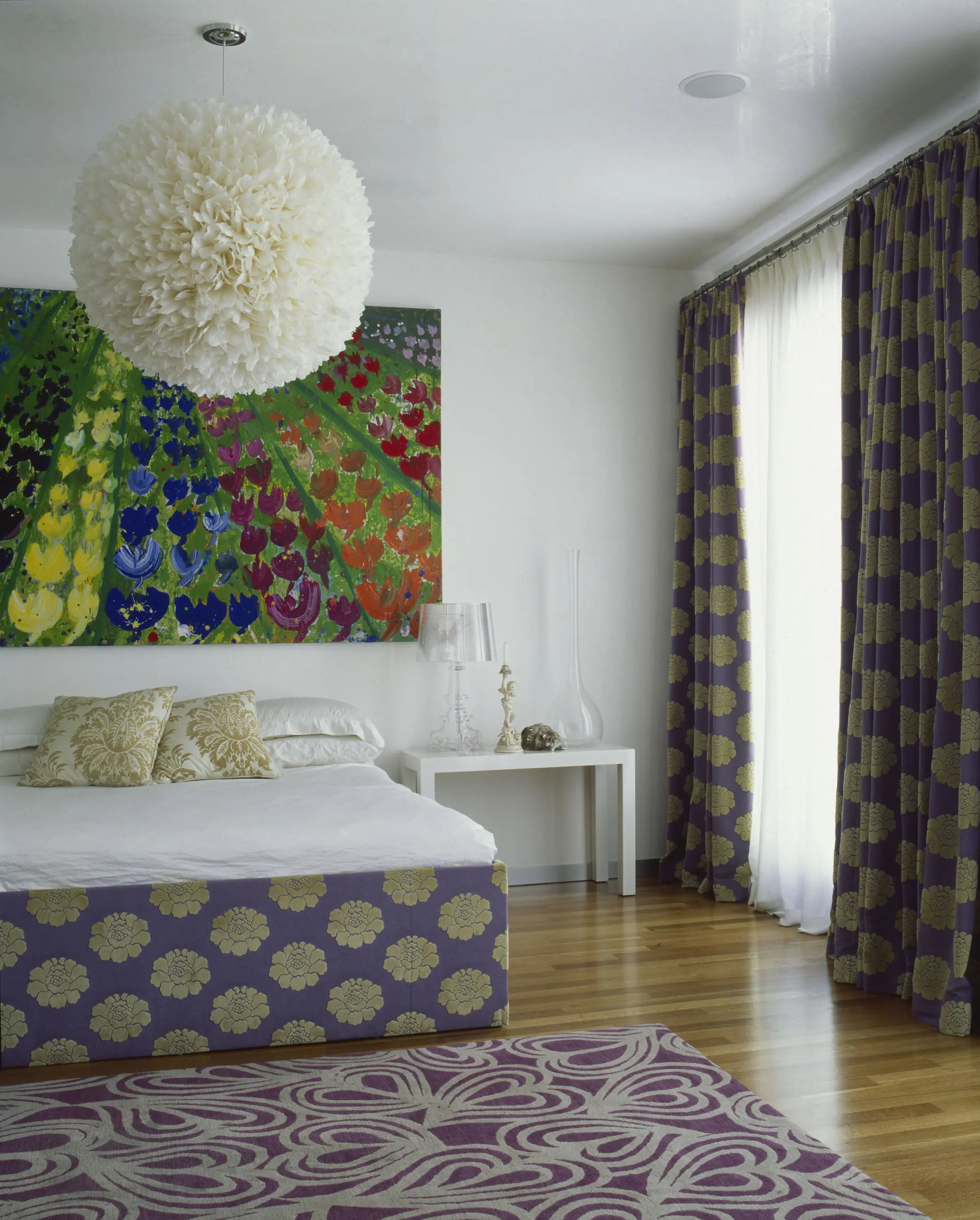 A bedroom with a bed featuring purple and gold floral bedding, a large fluffy white ceiling light fixture, and a vibrant floral painting on the wall. The room has a white nightstand with a glass vase and patterned purple curtains. A purple rug with a swirl pattern covers the wooden floor.