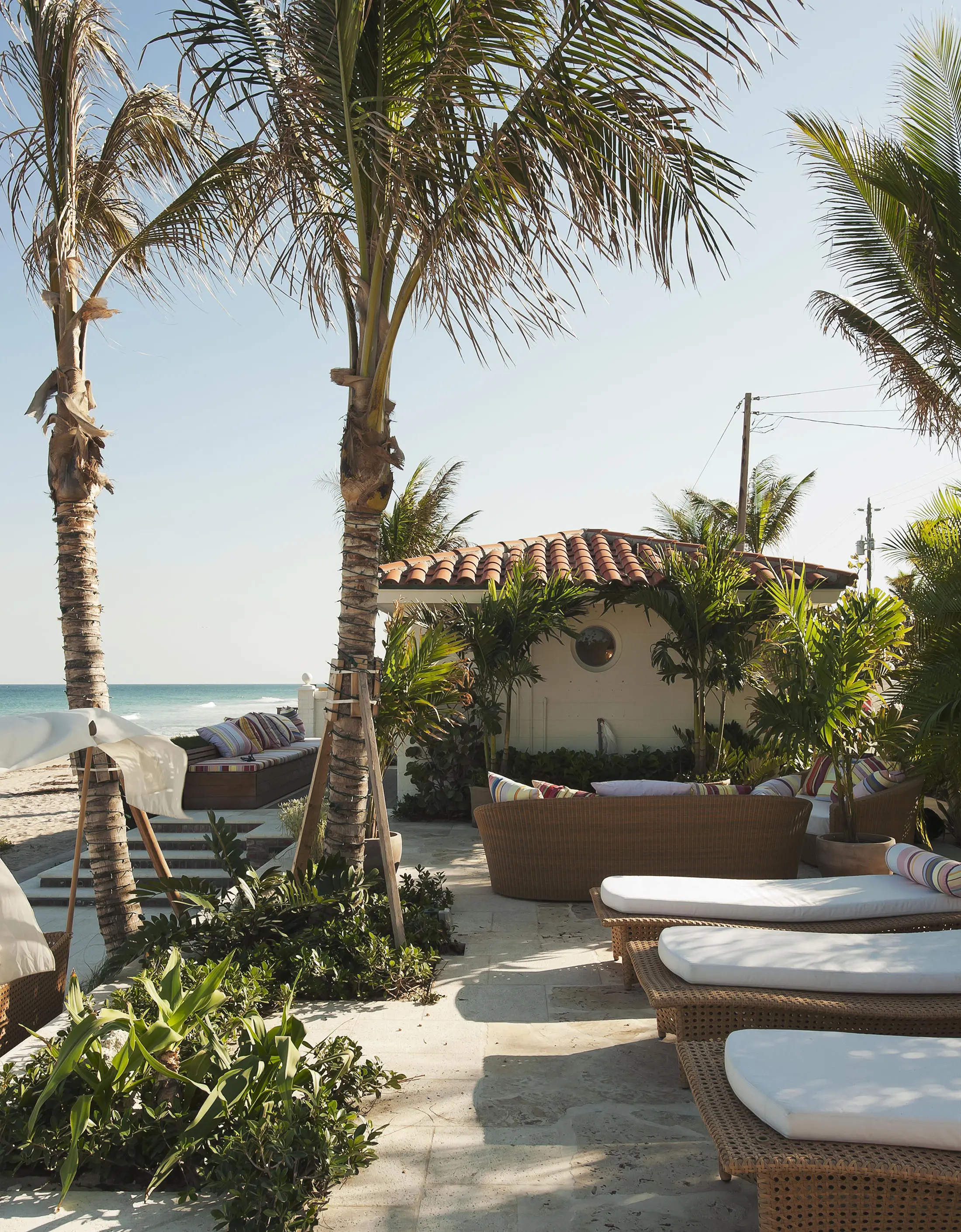 Tropical beachfront scene with palm trees and wicker lounge chairs on a stone patio. A small building with a red-tiled roof is surrounded by lush greenery. The ocean is visible in the background under a clear blue sky.