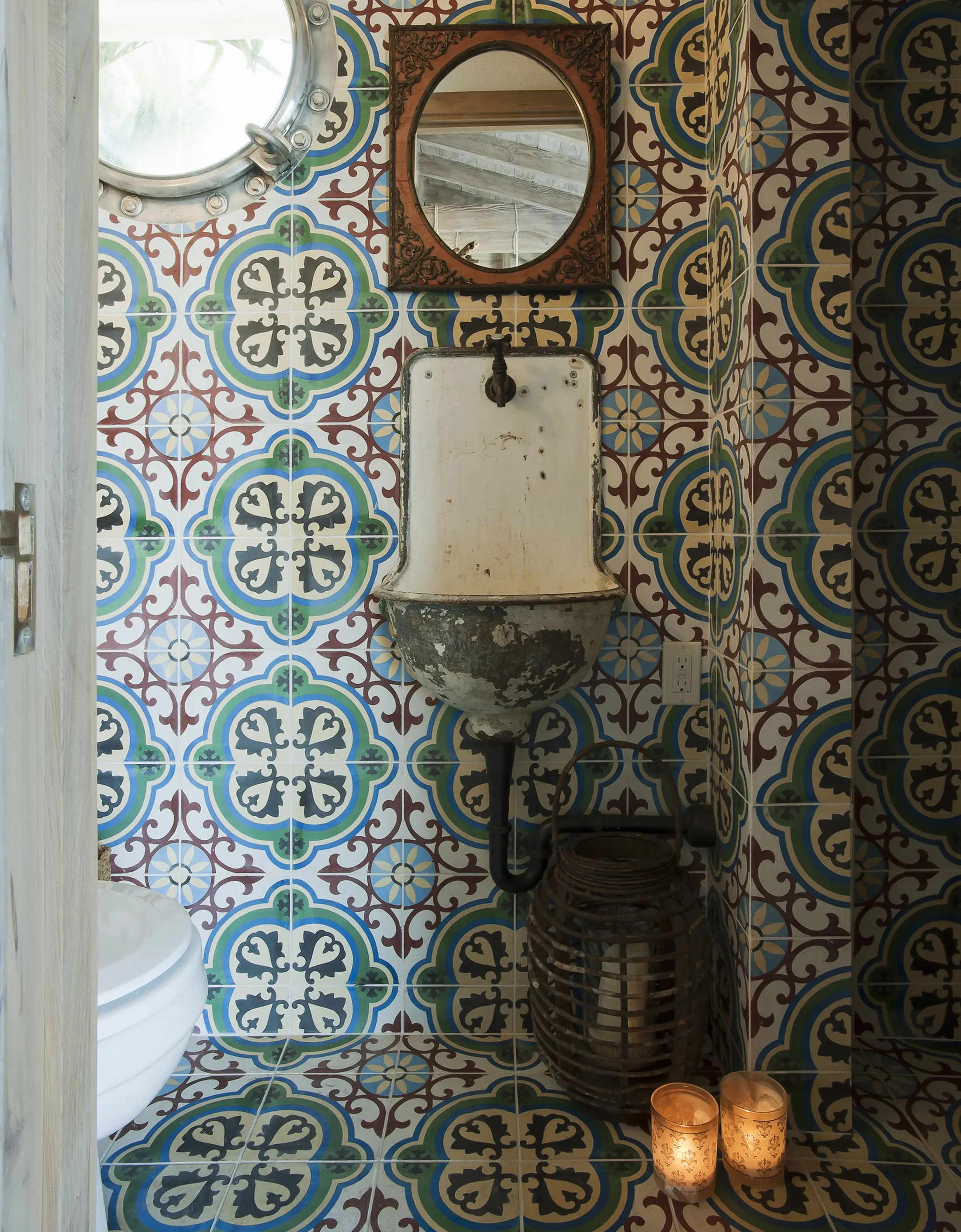 A small bathroom features vintage-patterned tiles in blue, red, and white. A rustic, wall-mounted sink is below an ornate, circular mirror. Two lit candles and a decorative basket sit on the floor. A round, porthole-style window adds natural light from the left.