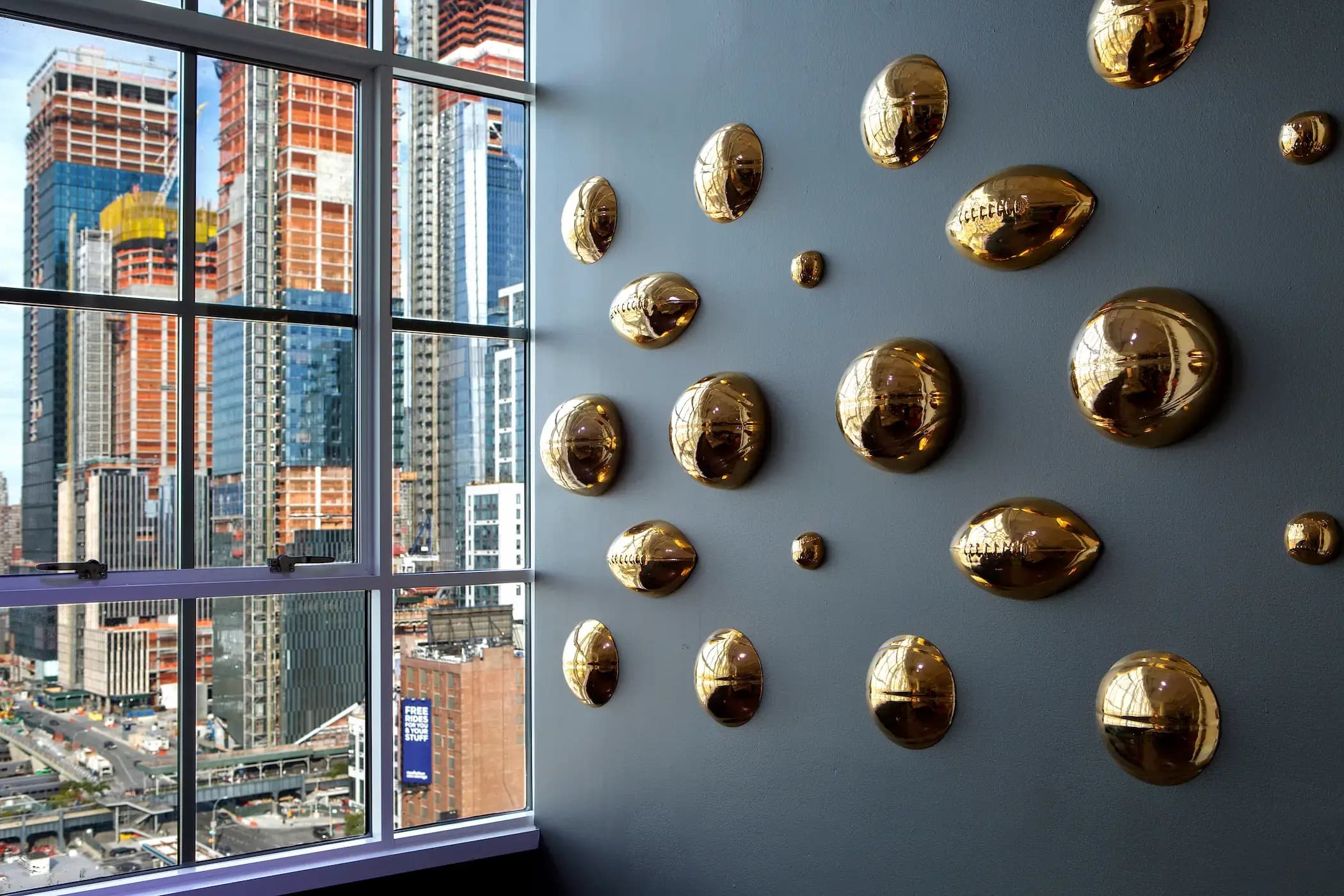 Wall with multiple gold, dome-shaped sculptures on the right. Large window on the left reveals a cityscape with numerous tall, modern skyscrapers under construction. Blue sky and scaffolding visible outside, creating a contrast between the indoor art and outdoor urban development.