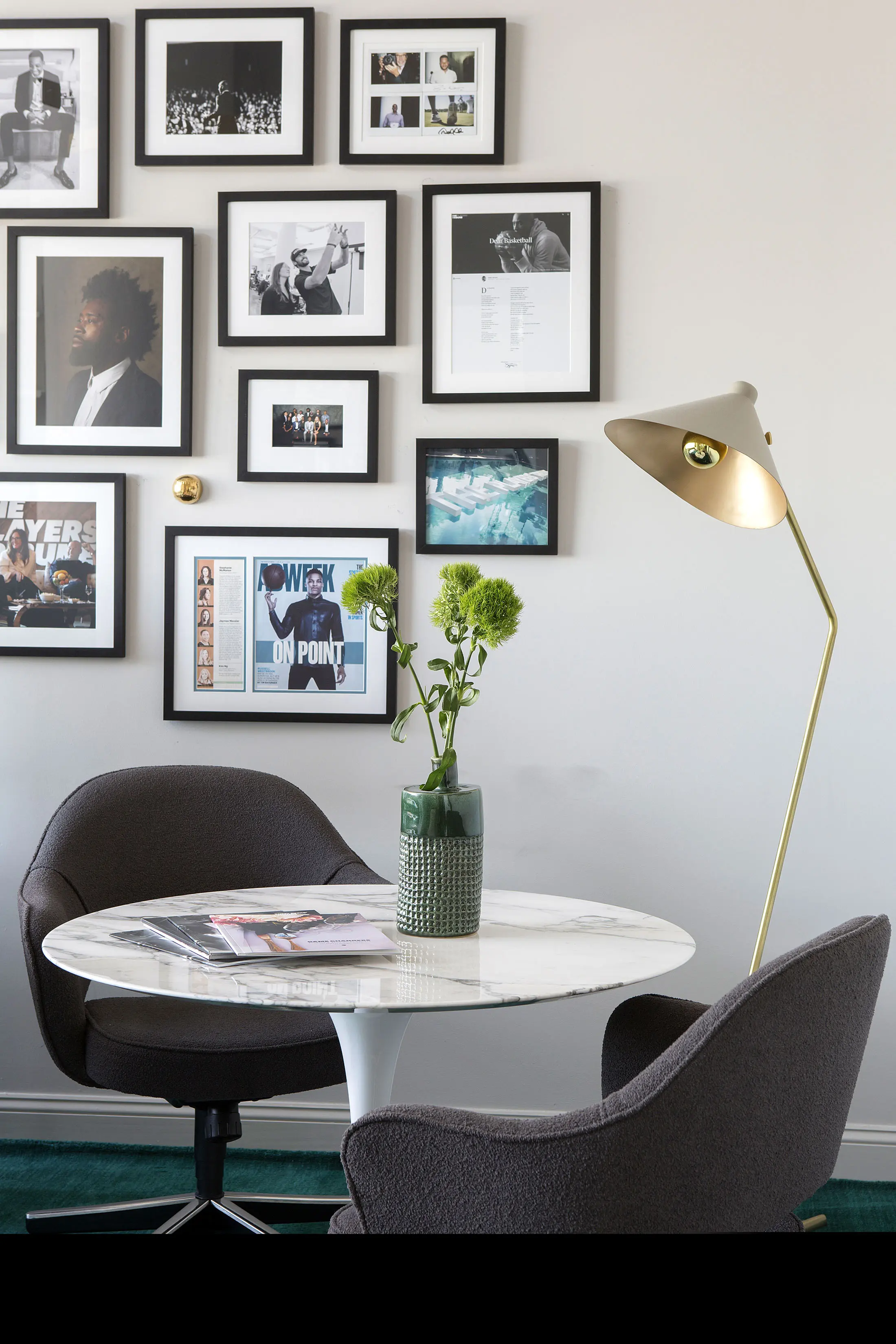 A stylish room with a round marble table, two dark grey chairs, and a textured green vase with flowers. A gold floor lamp stands nearby. A gallery wall displays various framed photographs and artwork in black frames against a light-colored wall. Magazines rest on the table.